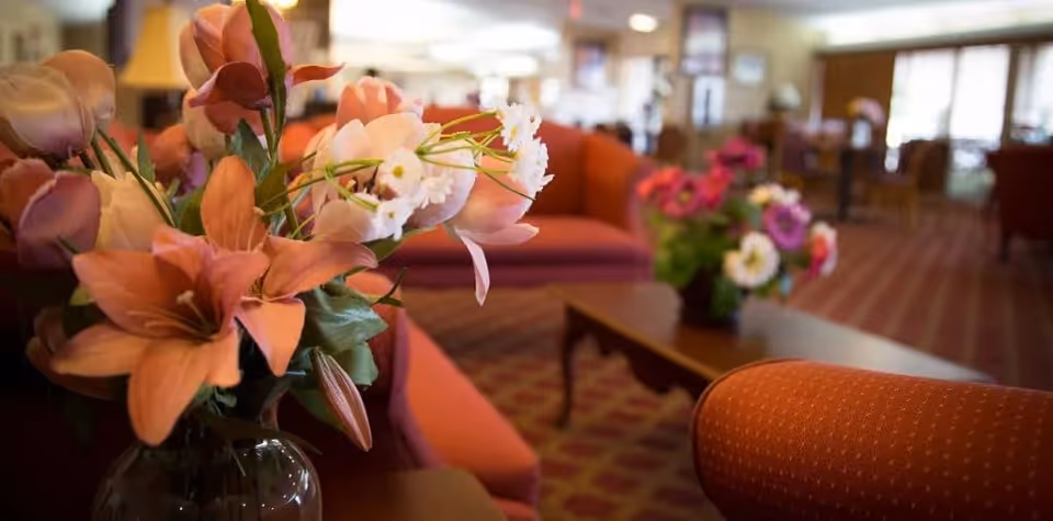 Close-up of a vase with pink and white flowers on a table in a cozy living room area with red upholstered chairs and sofas, a patterned carpet, and soft lighting.