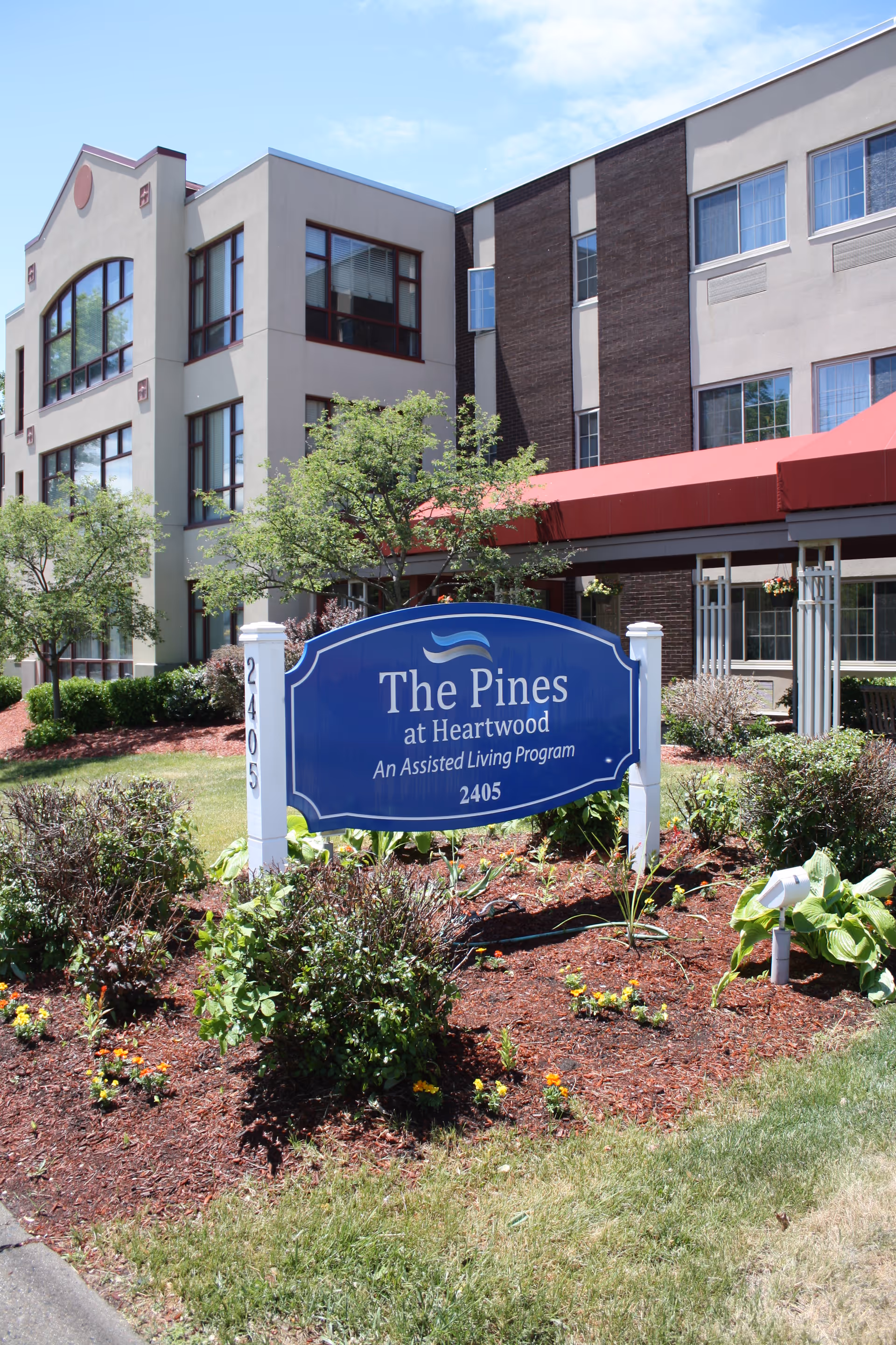 Front exterior of The Pines at Heartwood assisted living facility with a blue entrance sign amid landscaping.