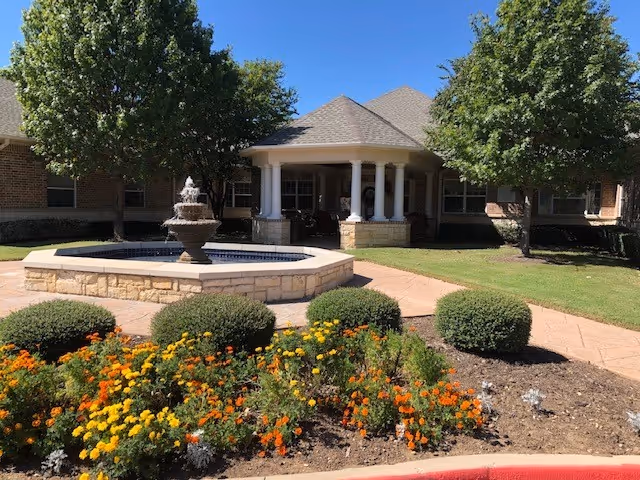 Outdoor view of Magnolia Assisted Living and Memory Care facility showing a landscaped garden with colorful flowers and bushes in front of a stone fountain. The building has a covered porch with white columns and a shingled roof, surrounded by green trees and a clear blue sky.