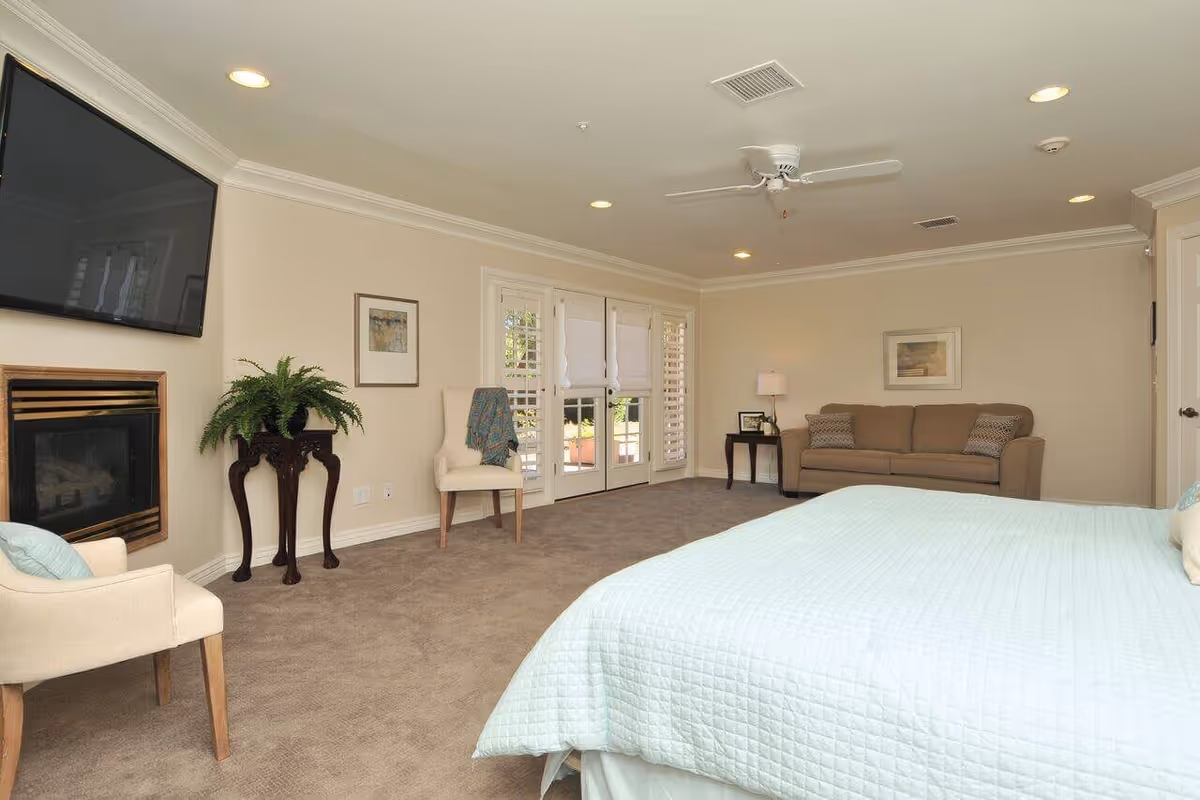 A spacious bedroom with a large bed covered in a light blue quilt. The room features beige walls, a ceiling fan, recessed lighting, a wall-mounted flat-screen TV above a fireplace, two beige chairs, a small table with a lamp, a brown sofa, and French doors with white blinds leading outside.