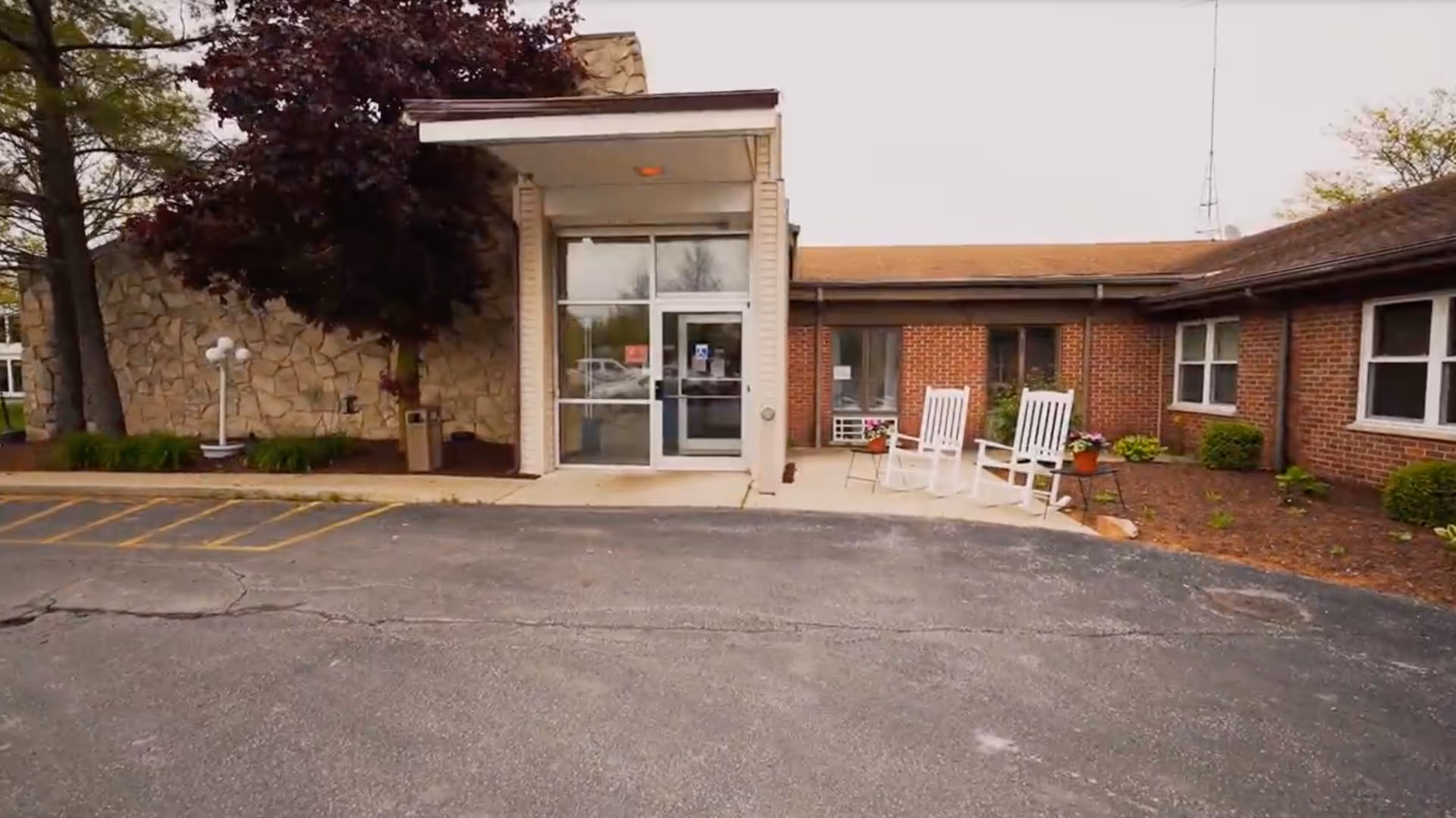 Front entrance of a single-story brick care facility with a covered glass doorway, two white rocking chairs on the porch, and a parking area in front.