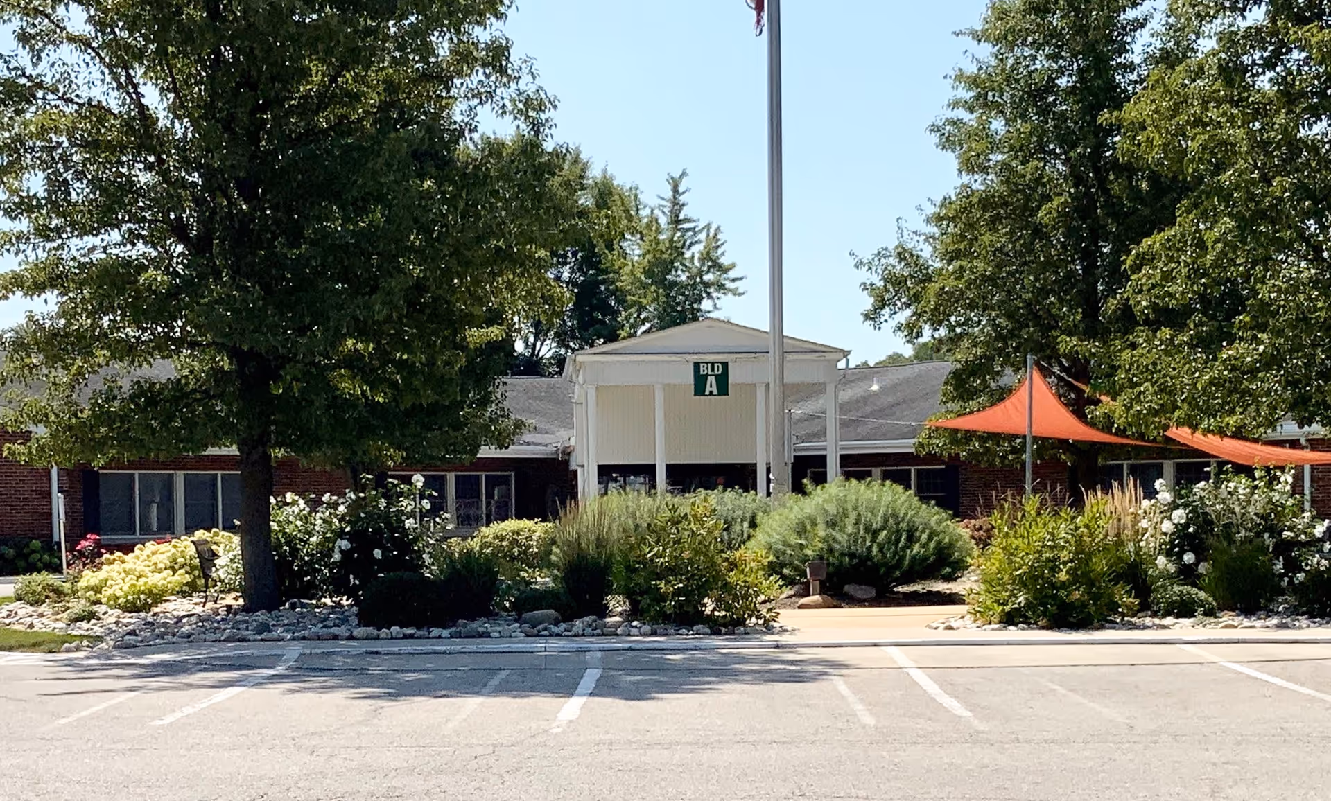 Exterior view of a single-story brick building labeled BLD A at Grace Village Retirement Community, surrounded by trees, bushes, and landscaped garden beds with a flagpole and orange shade sails in front.