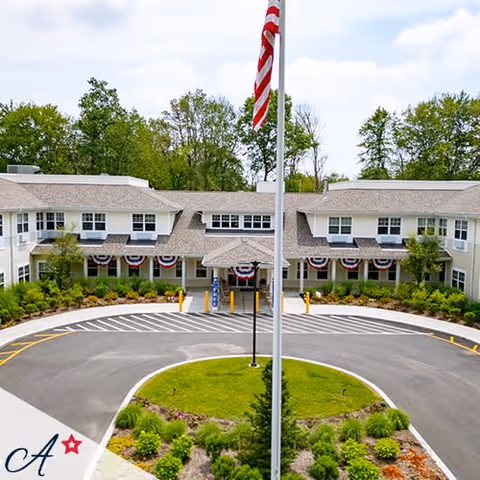 Front exterior view of All American Assisted Living At Enfield, showing a two-story building with a circular driveway, landscaped greenery, and an American flagpole in the center.