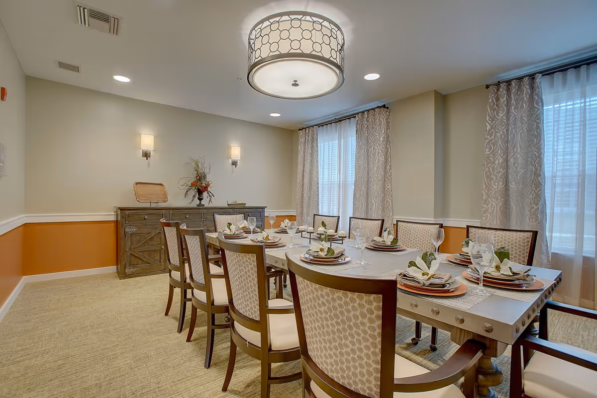 A well-lit dining room with a long rectangular table set for eight people. The table is decorated with neatly arranged plates, napkins with flowers, and wine glasses. The room features beige walls with an orange accent below the chair rail, patterned curtains on large windows, a wooden sideboard with a floral arrangement, and a modern ceiling light fixture.