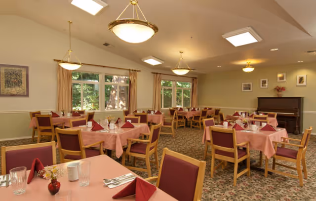 A senior-living dining room with several tables covered in pink tablecloths set with napkins, glassware and chairs beneath ceiling lights and windows.
