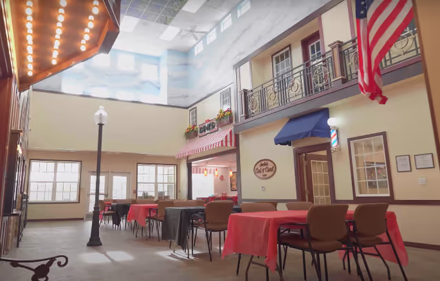Interior view of a senior living facility dining area designed to resemble a small town street with tables covered in red and gray tablecloths, chairs, a street lamp, and storefront facades including a diner and a barber shop. The ceiling is painted with a sky mural and there is an American flag hanging on the right side.