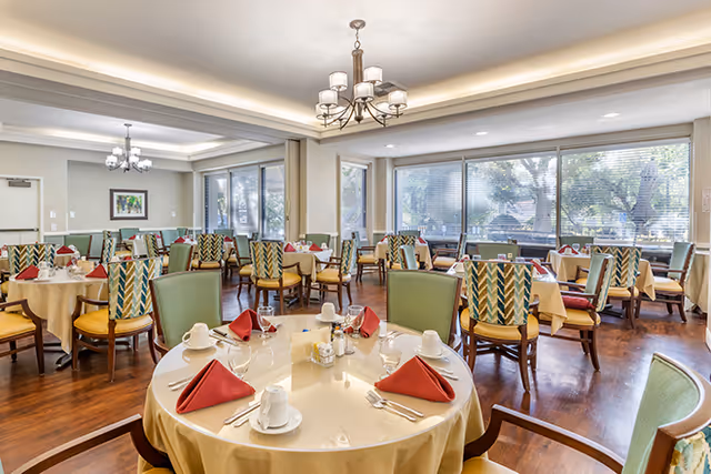 Dining room with round tables set for meals, upholstered chairs, chandeliers, and large windows overlooking trees.