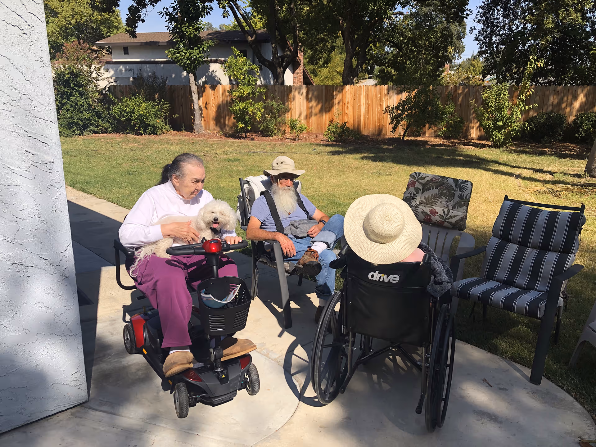 Three elderly individuals outdoors in a garden area. One woman is sitting on a mobility scooter holding a small white dog, a man with a long white beard and hat is sitting on a chair, and another person wearing a large sunhat is seated in a wheelchair. There are additional empty chairs nearby and a wooden fence with trees in the background.