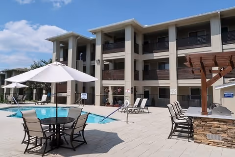 Outdoor pool area at MontClair Estates with lounge chairs, tables with umbrellas, and a pergola with a built-in grill, adjacent to a three-story residential building under a partly cloudy sky.