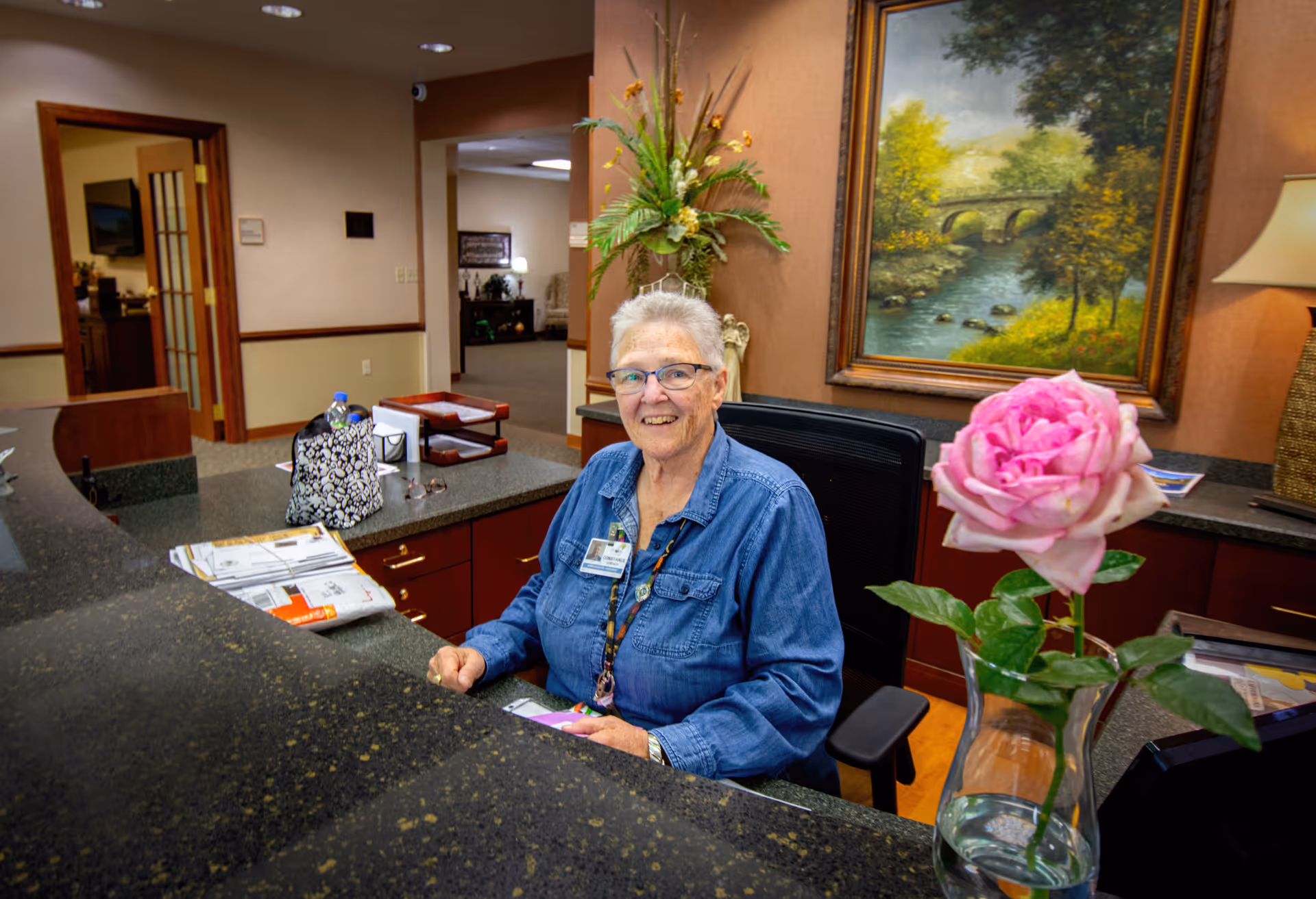 An elderly woman with short gray hair and glasses sits behind a reception desk in a senior care facility. She is smiling and wearing a blue denim shirt with a name badge. On the desk in front of her are papers, a patterned bag, and a vase with a pink rose. Behind her is a framed painting of a river and bridge, a tall floral arrangement, and a lamp.