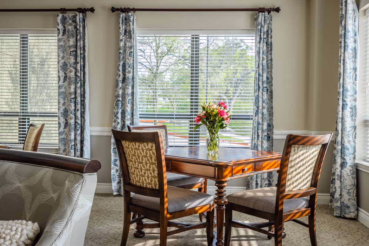 Sunlit dining area with a wooden table and four chairs by large windows with patterned curtains and a vase of flowers.