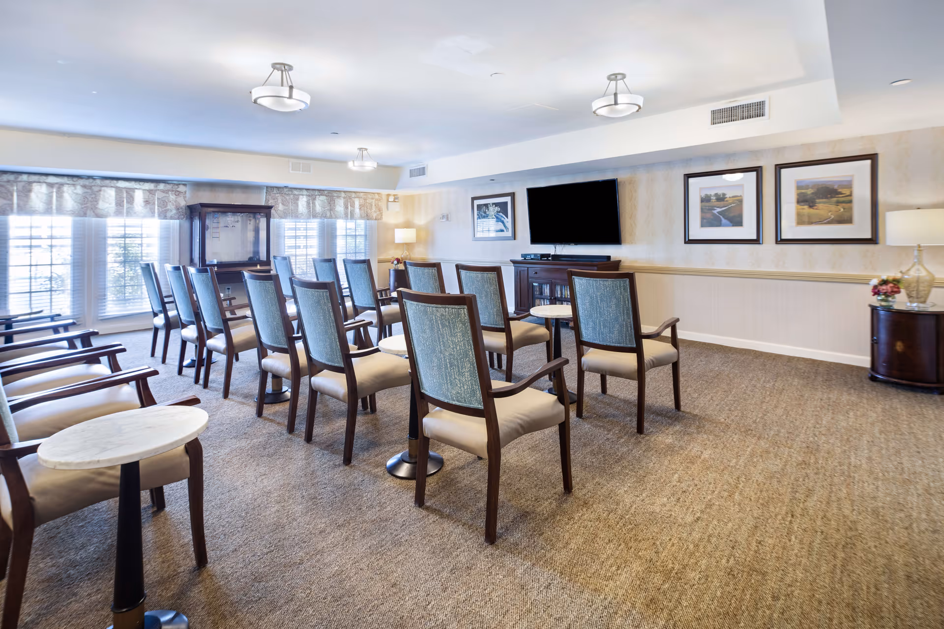 A well-lit community room with rows of wooden chairs featuring light blue upholstery, arranged facing a large flat-screen TV mounted on the wall. The room has beige carpet, light-colored walls with framed landscape pictures, and large windows with valances allowing natural light to enter. Small round tables are placed between some chairs, and there are lamps and a cabinet along the walls.