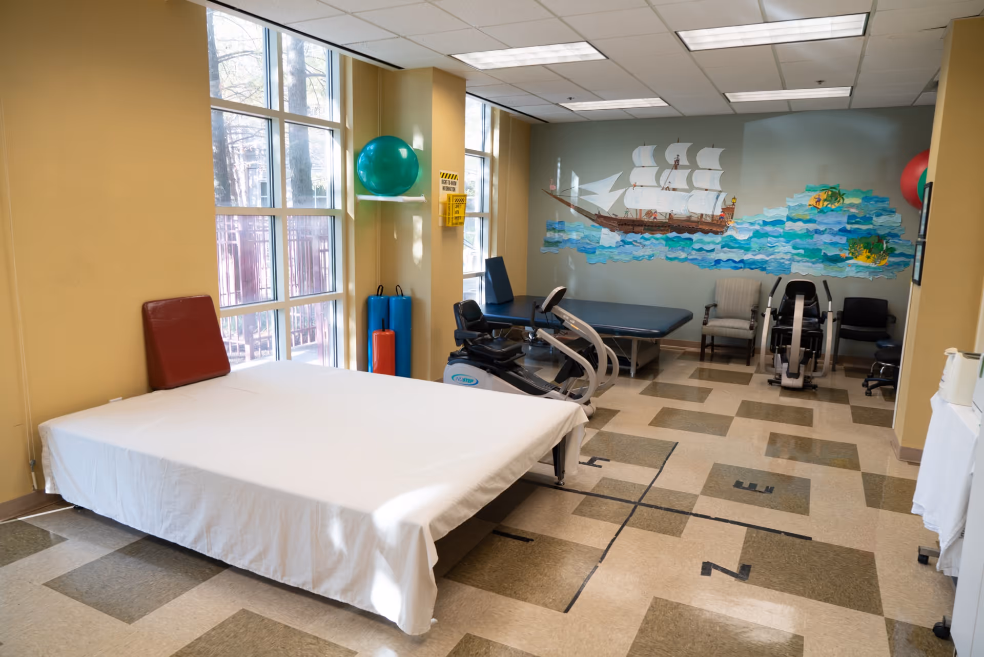 A physical therapy room with a large treatment table covered with a white sheet, exercise equipment including a stationary bike and rowing machine, chairs, and a colorful mural of a sailing ship on the wall. The room has large windows letting in natural light and exercise balls mounted on the walls.