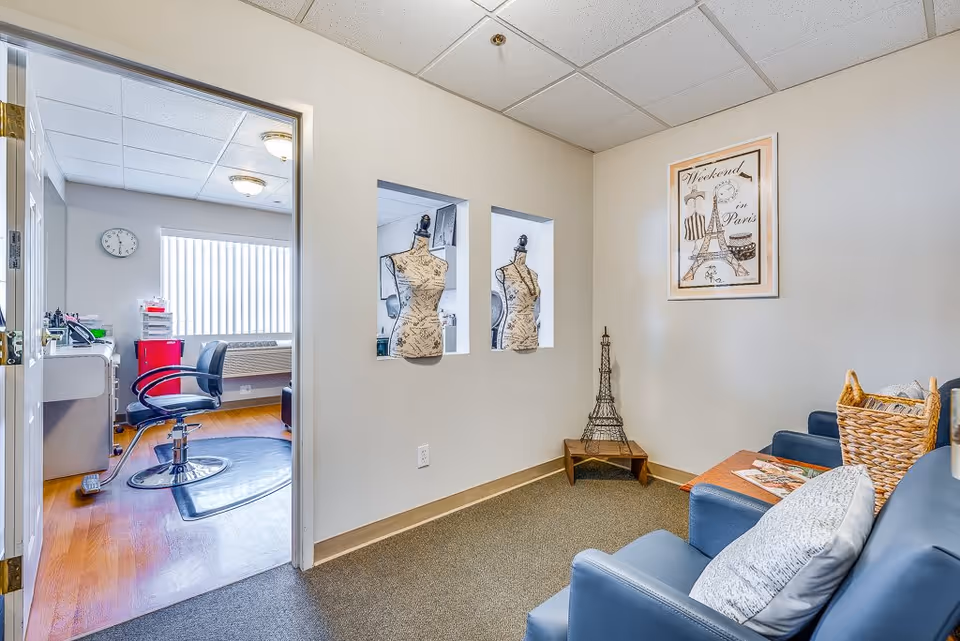 A cozy interior room with blue armchairs and a small wooden table holding a basket and magazines. The wall features a framed picture of the Eiffel Tower and two mannequin busts displayed in recessed wall niches. Through an open door, a bright room with a black swivel chair, a desk, and a clock on the wall is visible.
