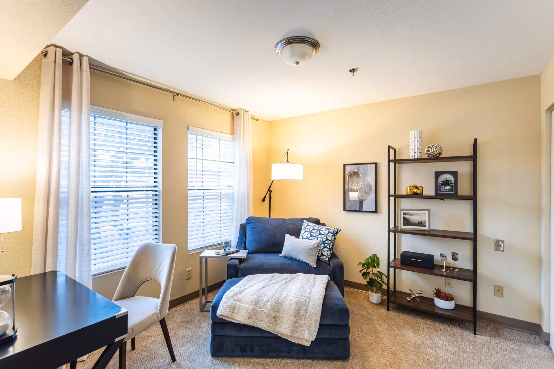 A cozy living room area with a blue armchair and matching ottoman covered with a beige throw blanket. There is a small side table with a glass of water and a book next to the chair. A floor lamp provides light, and two windows with white blinds and light curtains let in natural light. A black desk with a white chair is partially visible on the left. On the right side, there is a black metal and wood shelving unit with decorative items and framed pictures. The walls are painted a soft beige color.