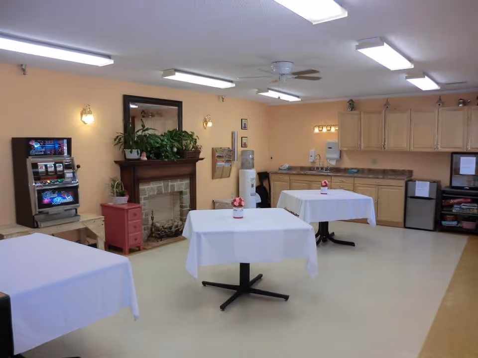 A common room with three tables covered in white tablecloths, each with a small flower arrangement. The room has a fireplace with plants on the mantle, a slot machine, a water cooler, and a kitchenette with cabinets, a sink, and a small refrigerator. The walls are painted peach, and the ceiling has fluorescent lights and a ceiling fan.