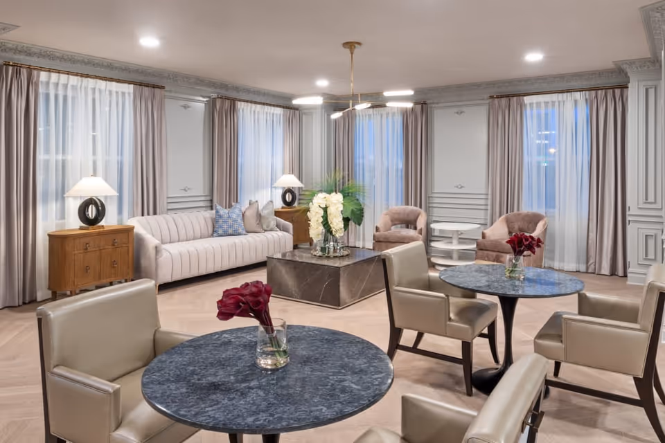 A spacious and elegant living room area with a light beige sofa, two armchairs, and two round tables with four beige chairs. The room features large windows with sheer white curtains and taupe drapes, wooden side tables with modern lamps, a square marble coffee table with a floral arrangement, and a modern chandelier hanging from the ceiling.