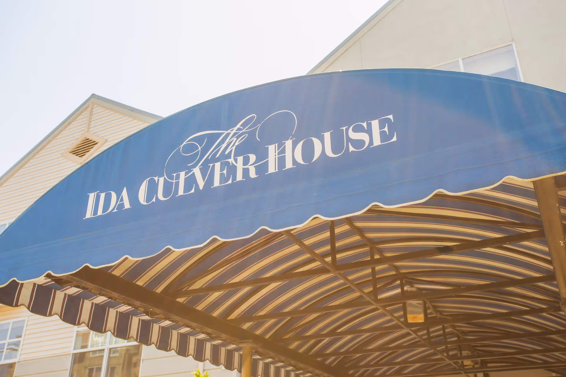 Blue and white striped awning with the text 'The Ida Culver House' in white letters, attached to a building with beige siding and windows visible in the background.