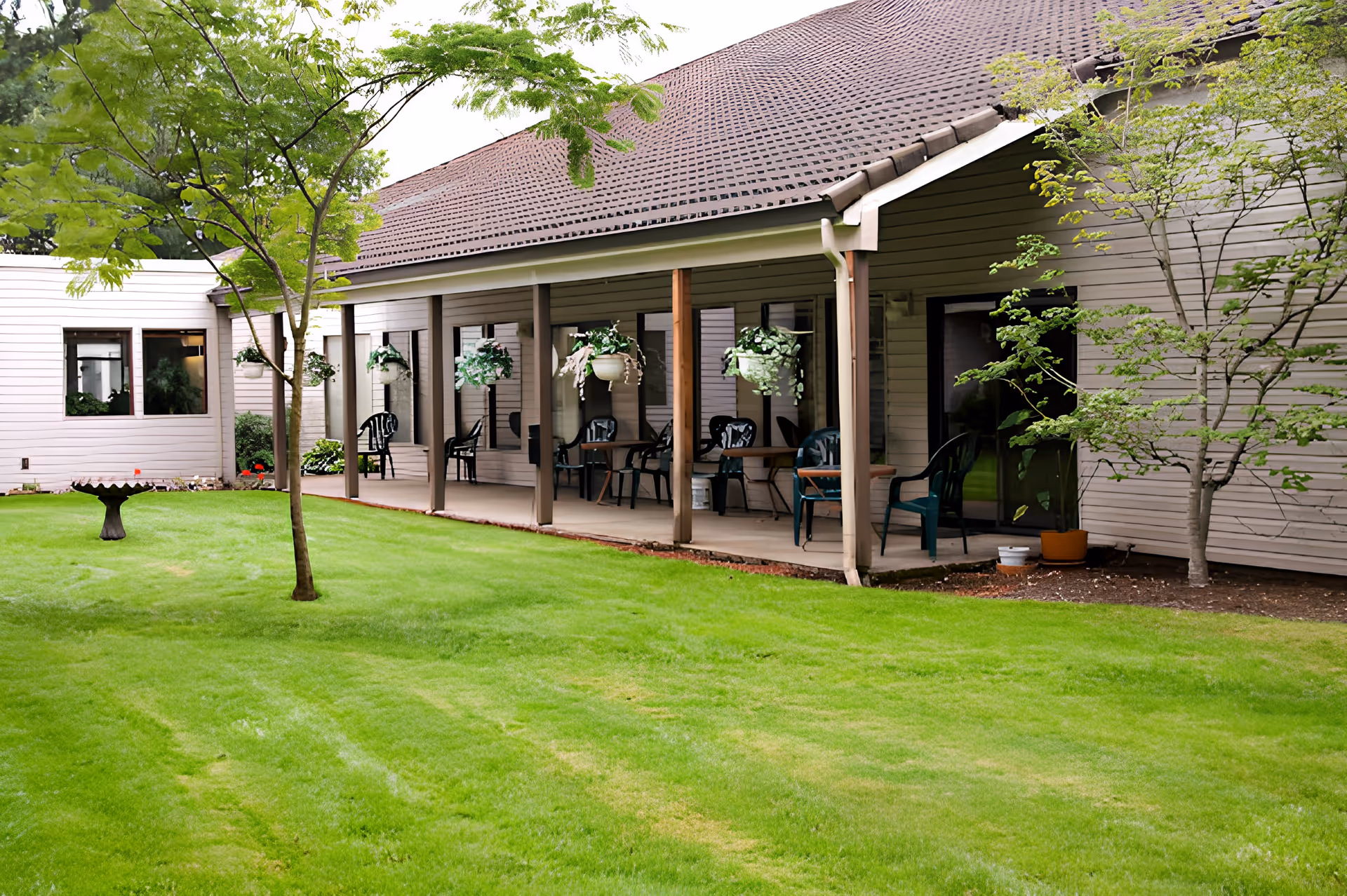A peaceful outdoor courtyard at Churchill Estates Retirement Community featuring a well-maintained green lawn, a birdbath, several small trees, and a covered patio with hanging flower pots and plastic chairs arranged around small tables.