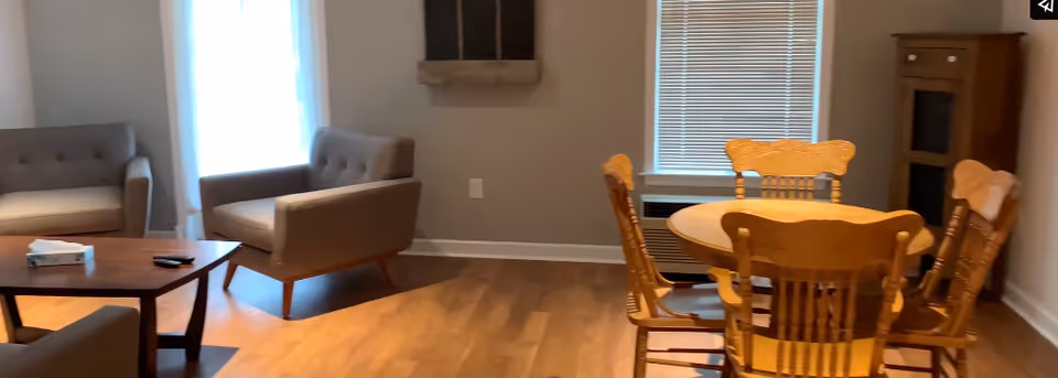 A cozy living and dining area with wooden flooring. The living area has two gray upholstered armchairs and a matching loveseat around a wooden coffee table with a tissue box and remote control on it. The dining area features a round wooden table with four matching chairs. There is a window with blinds and a wooden cabinet in the corner.