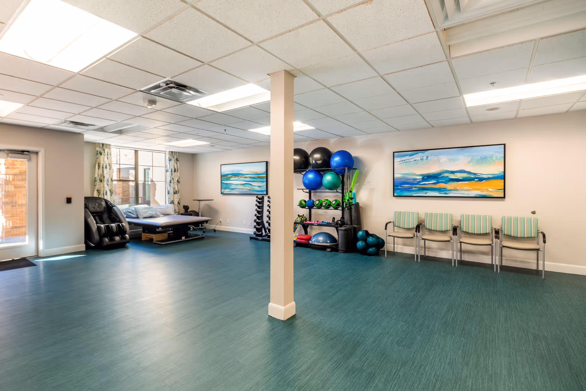 A spacious room with teal flooring and beige walls featuring exercise equipment including stability balls, dumbbells, and a massage chair. There are four chairs lined up against the wall under a colorful landscape painting, and a large window with floral curtains letting in natural light.
