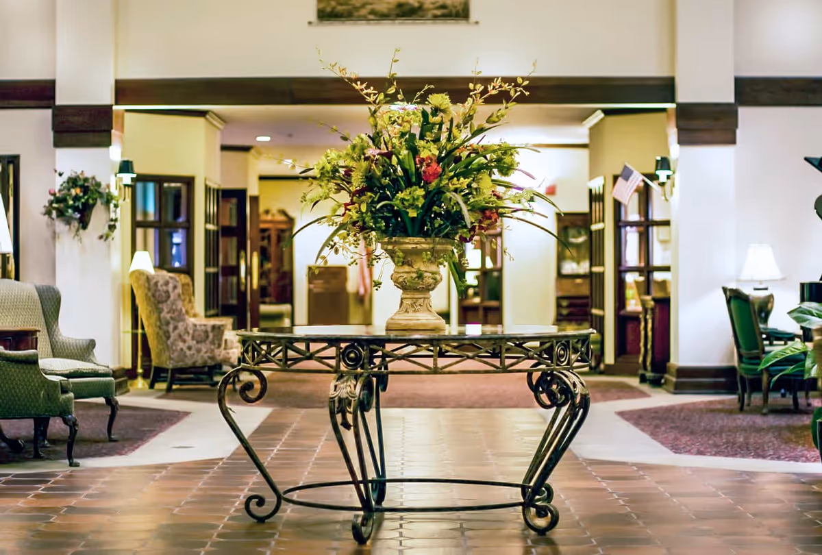 A spacious and elegantly decorated senior living facility lobby with a large floral arrangement on a wrought iron table in the center. The area features comfortable armchairs, warm lighting, and a tiled floor with carpeted sections.