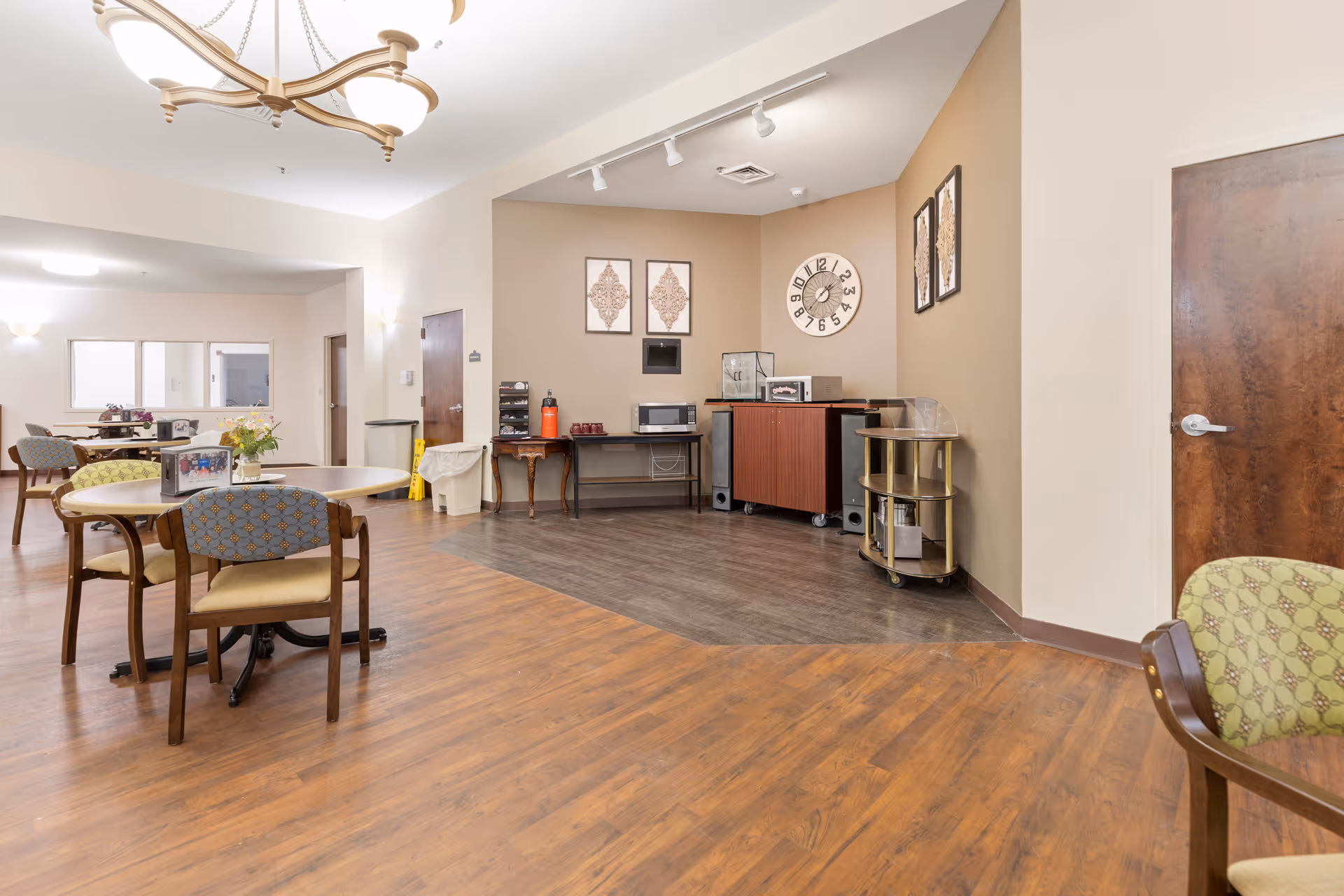 A spacious common area in a senior living facility with wooden flooring and beige walls. The room features round tables with chairs, a chandelier, wall art, a large clock, and a small kitchenette area with a microwave, coffee maker, and other appliances. There are doors and windows visible in the background.
