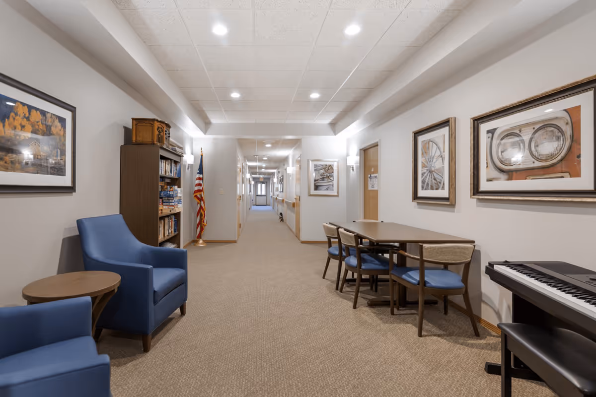 A well-lit hallway in a senior living facility with beige walls and carpet. On the left side, there are two blue armchairs and a wooden bookshelf filled with books and board games. An American flag stands next to the bookshelf. On the right side, there is a table with four chairs and a keyboard piano with a bench. The walls are decorated with framed pictures, and several doors line the hallway.