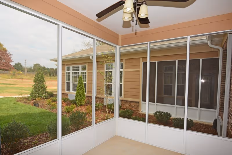 View from a screened-in porch showing a ceiling fan with lights, looking out onto a landscaped garden area with shrubs and small trees, and the exterior walls and windows of the Webb House Retirement Center building.