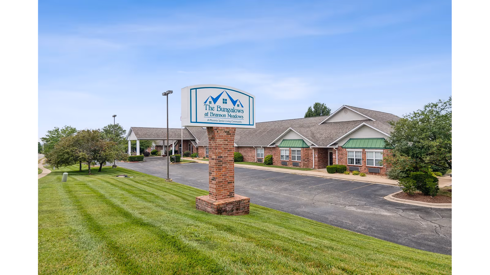 Exterior view of The Bungalows at Branson Meadows senior living community showing a brick signpost with the facility name, a paved driveway, well-maintained green lawn, trees, and a single-story brick building with green awnings under a clear blue sky.