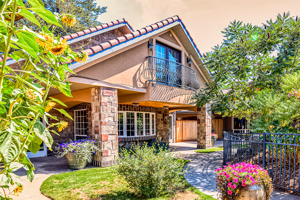 Exterior view of a senior living facility building with stone pillars, a balcony with wrought iron railing, and a tiled roof. The foreground features a curved walkway surrounded by green grass, colorful flowers, and various plants and trees under a partly cloudy sky.