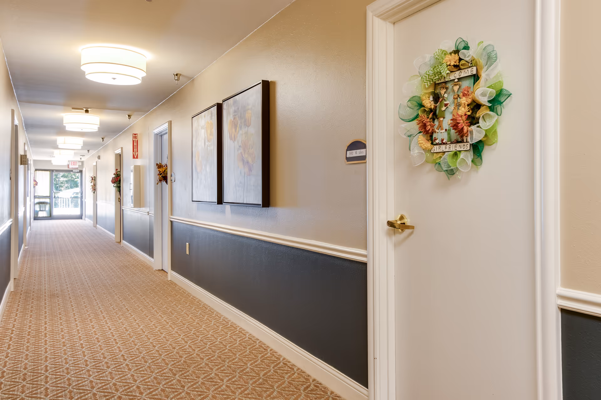 A long, well-lit hallway in an assisted living facility with beige walls and patterned carpet. The hallway features multiple doors, one of which has a decorative wreath with the words 'Welcome Girlfriends'. There are framed floral paintings on the walls and ceiling lights evenly spaced along the corridor. At the end of the hallway, there is a glass door leading outside.