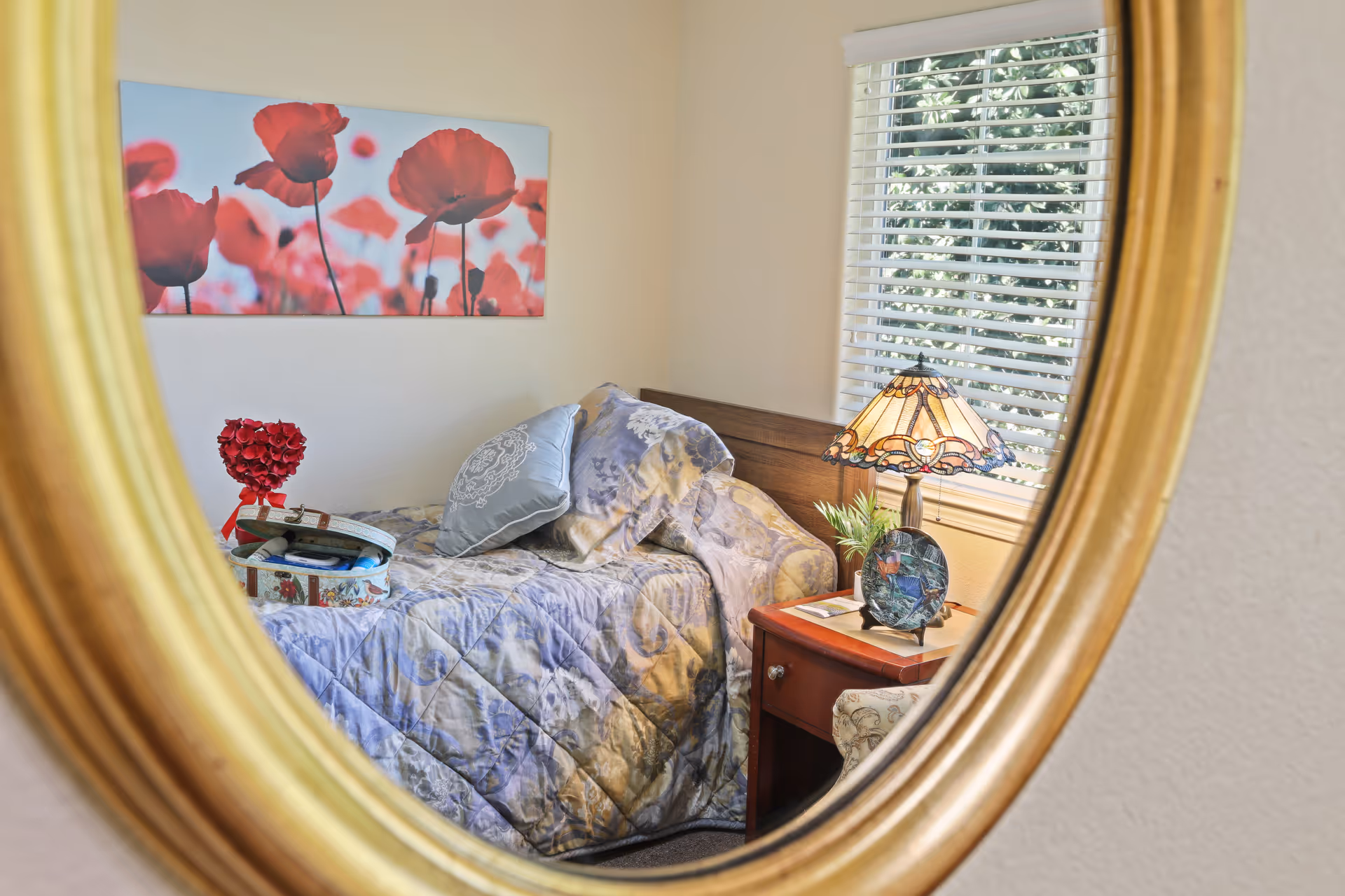 Reflection in a round mirror showing a cozy bedroom with a bed covered in a patterned quilt and pillows, a bedside table with a decorative lamp and a small plant, a window with white blinds, and a wall art featuring red poppies.