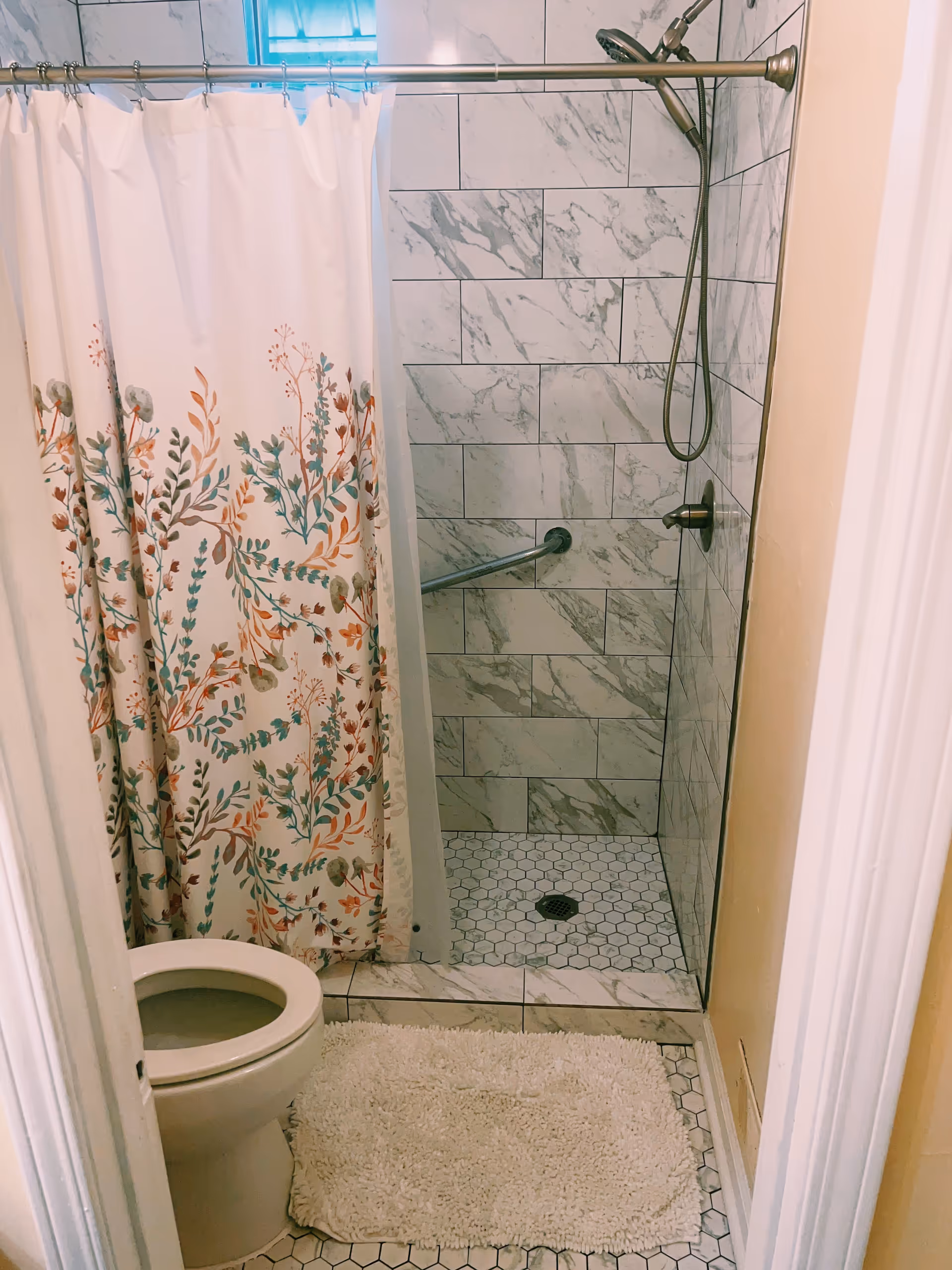 Small bathroom with a toilet, a white and floral shower curtain, a walk-in shower with marble-patterned tiles, a handheld showerhead, and a white bath mat on the hexagonal tiled floor.