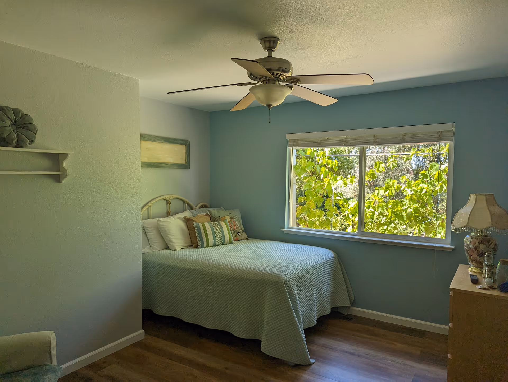 Sunlit bedroom with a bed against a blue wall, ceiling fan, window showing green foliage, and a dresser with a lamp.