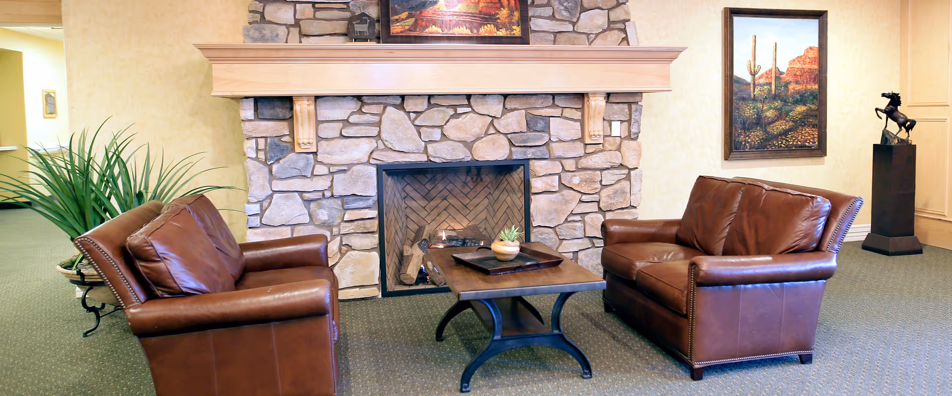 A cozy sitting area with two brown leather armchairs facing each other across a wooden coffee table. Behind the table is a stone fireplace with a wooden mantel. On the mantel is a framed picture, and another framed picture hangs on the wall to the right. A potted plant is placed to the left of the armchairs, and a horse statue on a pedestal is visible in the background.