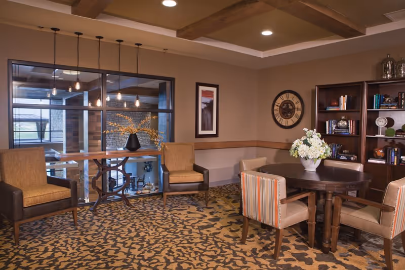 A cozy interior lounge area with a round wooden table surrounded by four upholstered chairs, a bookshelf filled with books and decorative items, two armchairs near a window with hanging pendant lights, a wall clock, and a framed picture on the wall. The room has a patterned carpet and wooden ceiling beams.