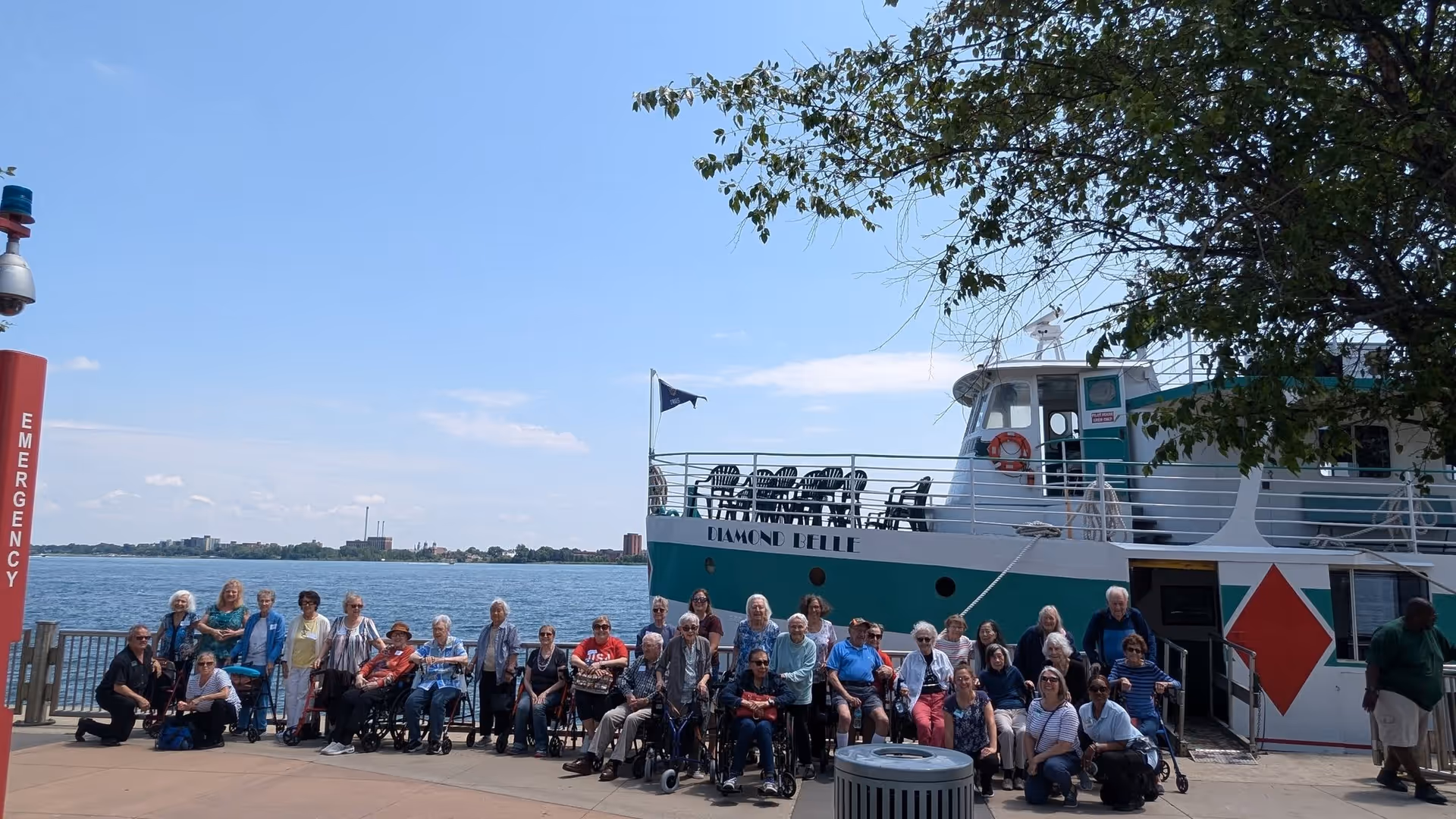 A large group of elderly people, some in wheelchairs, posing for a photo on a dock next to a large boat named Diamond Belle. The background shows a body of water and a clear sky with some clouds. There is a tree partially visible on the right side and an emergency call station on the left.
