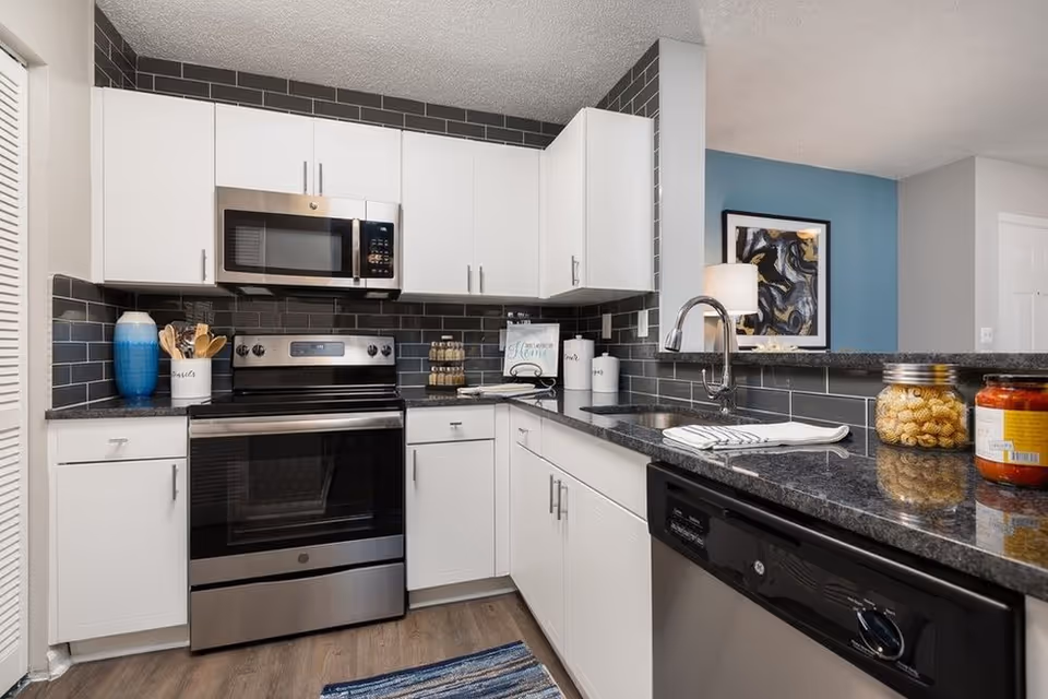 Modern kitchen with white cabinets, black granite countertops, stainless steel oven and microwave, black subway tile backsplash, a sink with a curved faucet, and various kitchen items including a blue vase, utensil holder, spice rack, and jars of pasta and sauce on the counter.