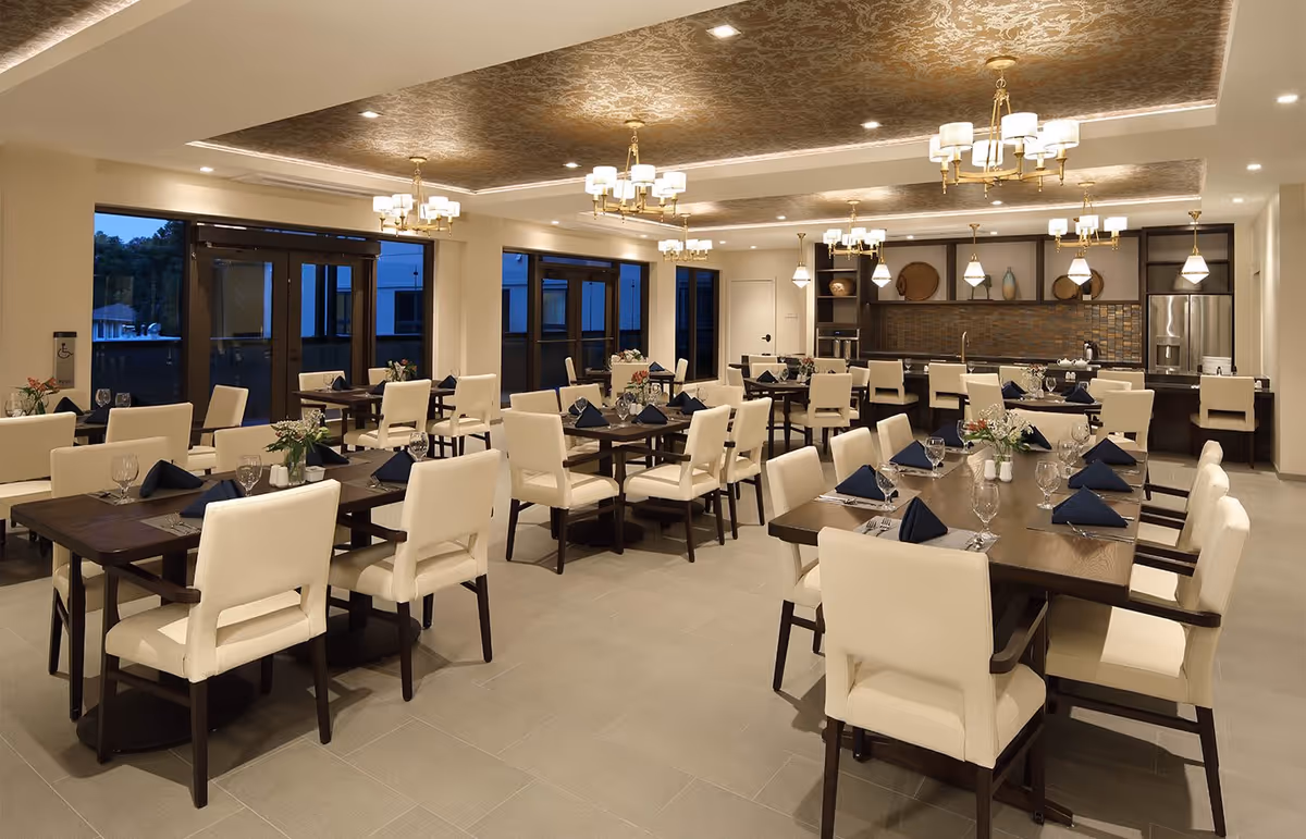 A spacious and elegant dining room with multiple dark wood tables and cream upholstered chairs. Each table is set with navy blue folded napkins, glassware, and silverware. The ceiling features decorative gold patterns and multiple chandeliers with white lampshades. Large windows and glass doors line one side of the room, and there is a kitchen area with a refrigerator and counter seating in the background.