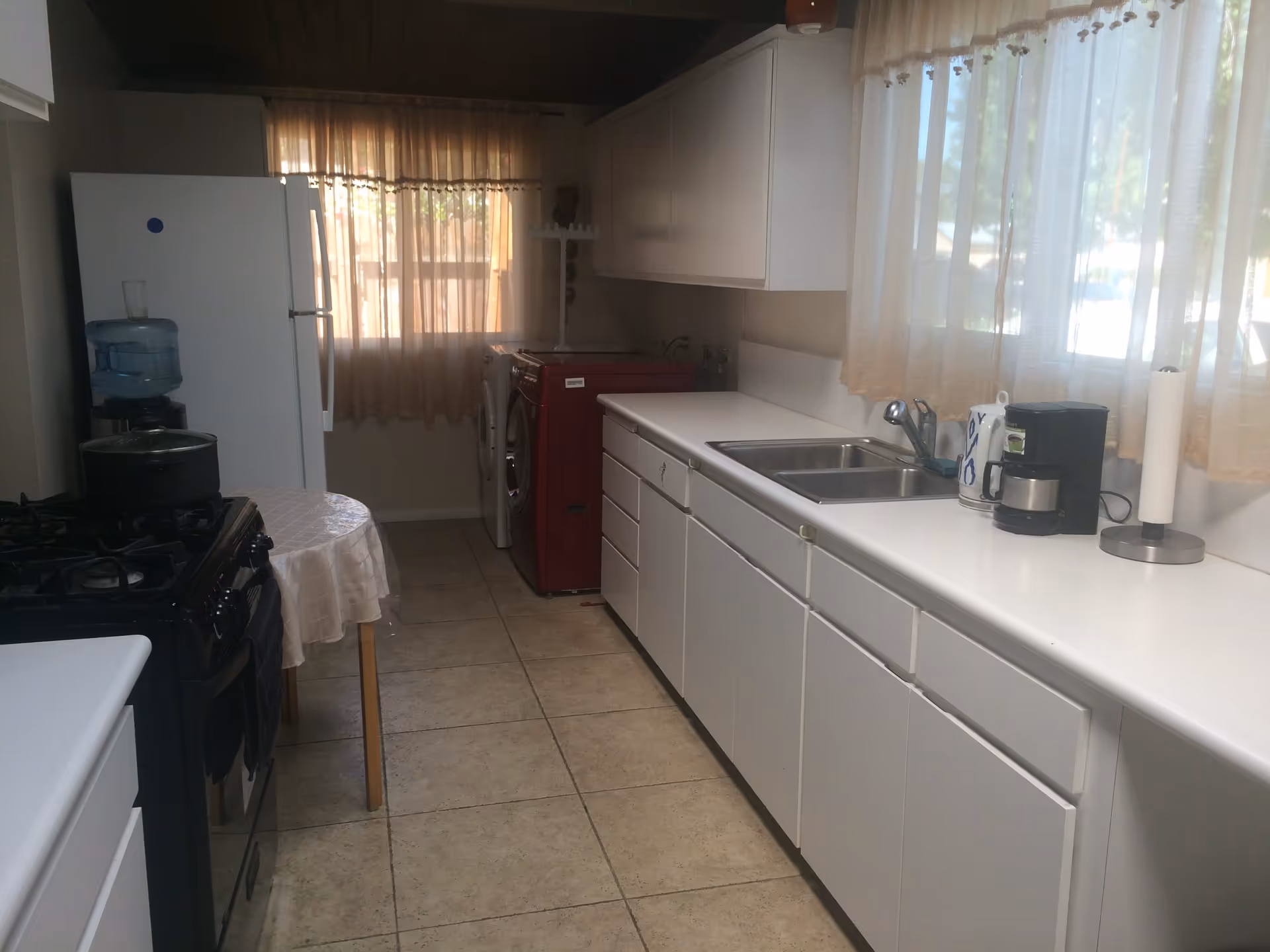 A narrow kitchen with white cabinets and countertops on the right side, a double sink, a coffee maker, and a paper towel holder. On the left side, there is a black stove with a pot on it, a small round table with a tablecloth, a water dispenser, and a white refrigerator. At the far end, there is a red washing machine and a white dryer under a window with sheer curtains.