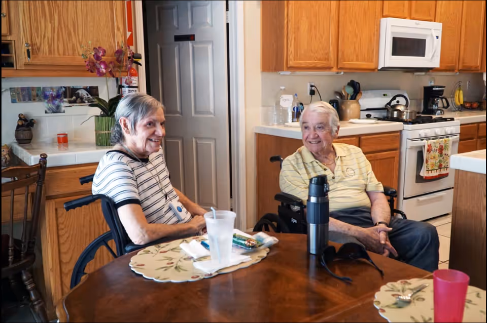Two elderly individuals in wheelchairs sitting at a wooden dining table in a kitchen. The kitchen has wooden cabinets, a white microwave, stove, and various kitchen items on the counters. One person is wearing a striped shirt and the other a yellow shirt. There are placemats, cups, and a thermos on the table.