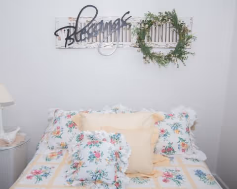 A cozy bedroom with a bed covered in floral-patterned bedding and pillows. Above the bed, there is a decorative wall piece with the word 'Blessings' and a green wreath. A small round side table with a lamp is visible on the left side.