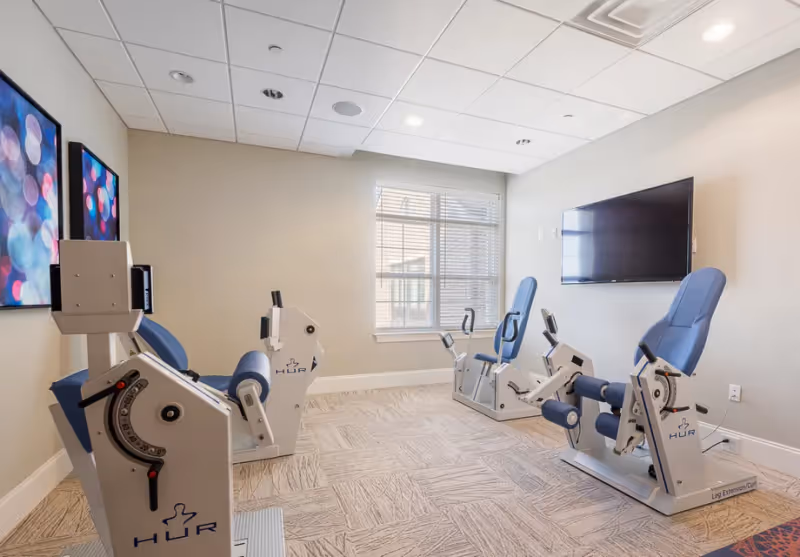 A small exercise room with three HUR brand seated exercise machines positioned facing a wall-mounted flat screen TV. The room has beige walls, a window with blinds, two colorful abstract paintings on one wall, and a patterned carpet floor.