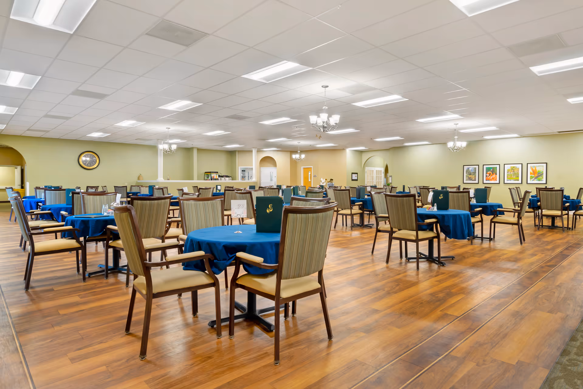 A spacious dining room with multiple round tables covered with blue tablecloths, each surrounded by several cushioned chairs. The room has wooden flooring, light green walls, chandeliers hanging from a white ceiling, and framed artwork on the far wall.