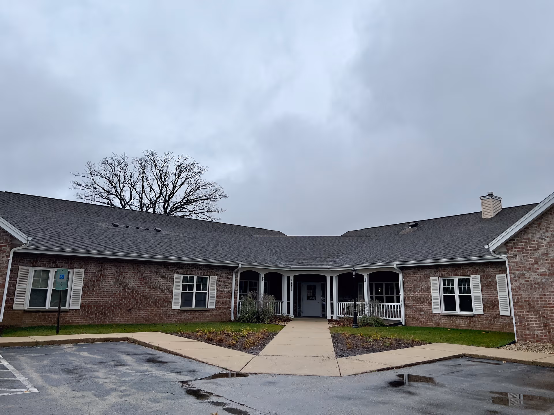 Front exterior view of a single-story brick building with a central entrance, white window shutters, and a covered porch area. The sky is overcast and there is a bare tree visible behind the building. A parking lot with some wet patches is in the foreground.