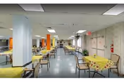 A spacious dining room with multiple tables covered in yellow floral tablecloths, each surrounded by chairs. The room has a tiled ceiling with fluorescent lighting, large windows on one side, and a light blue floor. There are columns and some plants near the windows.