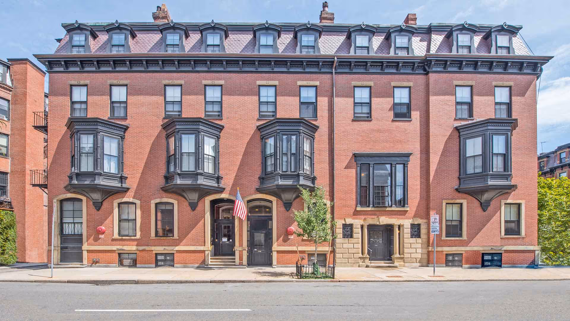 Front exterior view of a large, historic brick building with black framed windows and bay windows. The building has a classic architectural style with a mansard roof and dormer windows. There is an American flag displayed near the entrance and a small tree planted on the sidewalk in front.