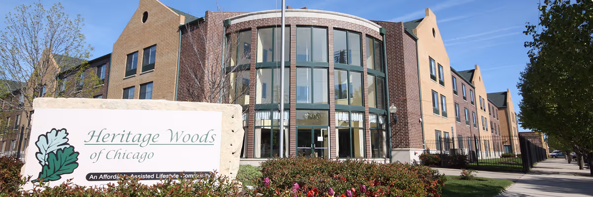 Exterior view of Heritage Woods of Chicago, a multi-story brick building with large windows and a landscaped front area with bushes and flowers. A sign in the foreground reads 'Heritage Woods of Chicago' with a green leaf logo.
