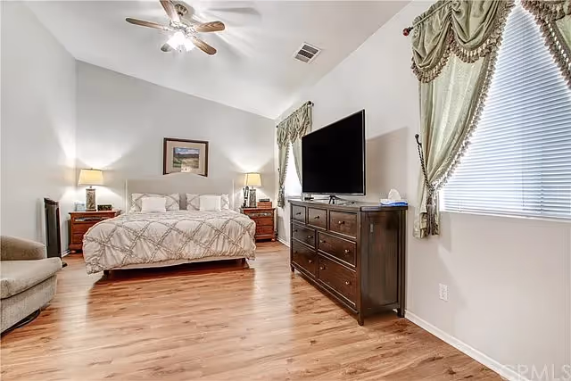 A spacious bedroom with a large bed featuring a patterned comforter, two bedside tables with lamps, a dresser with a flat-screen TV on top, a comfortable armchair, wood flooring, and windows with green curtains.